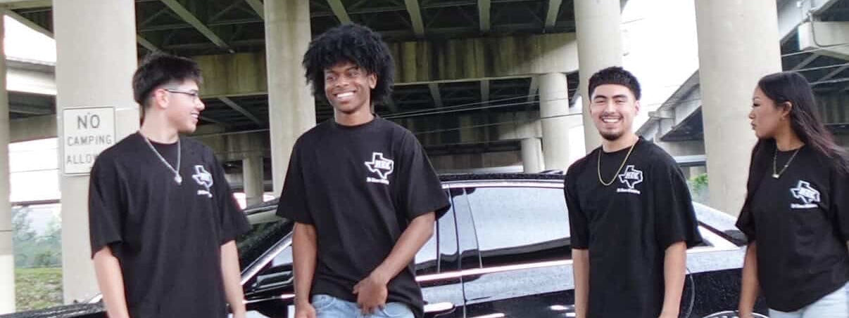 Four individuals wearing black t-shirts with a logo standing in front of a limousine.